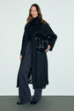A woman stands against a plain background, wearing an elegant manteau long femme in black, paired with a sleek black turtleneck, patterned wide-leg jeans, black shoes, and holding a stylish black handbag.