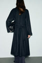 A person with long brown hair wears a manteau long femme, styled as a dark coat with a belt, holding a black handbag. The person is facing away from the camera against a plain light background.