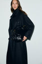 A woman in a long, dark trench anthracite coat stands against a light background, holding a large black leather handbag under her arm.