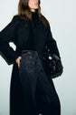 A woman in a black manteau long femme and patterned jeans stands against a light background, holding a black handbag at her side. Her face is partially out of frame.