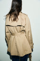 A person with medium-length brown hair, seen from behind, wearing a coton beige trench femme with a belted waist and dark pants, standing against a plain light-colored wall.