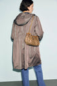 A person with short brown hair, wearing a parka mi-longue à capuche in light brown and blue jeans, stands facing away. They carry a brown, patterned shoulder bag with a gold chain strap.