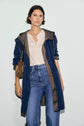 A woman in a navy blue parka réversible, light beige blouse, and high-waisted blue jeans stands against a plain background, holding a small brown purse with a gold chain strap.