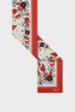 A cream-colored foulard femme with red borders and a floral pattern in red, blue, and brown. This carré rétro is folded in a zigzag shape, showing off the charming motif floral and a small logo in the corner.