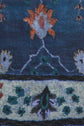 Close-up of an echarpe bleue, showcasing a blue fabric with a motif fleuri featuring stylized orange and teal floral motifs arranged in rows, separated by a thin black horizontal line, against a textured background.