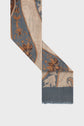 A close-up of an écharpe rectangulaire featuring a multicolor design with blue, beige, and brown floral and paisley patterns on fabric, shown with frayed edges against a white background. This elegant echarpe motif cachemire highlights the timeless blend of colors for a refined accessory.
