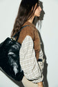 Woman in profile wearing a brown gilet sans manche cachemire over a plaid shirt, beige pants, and carrying a large black leather bag on her shoulder, standing against a white wall with a shadow.