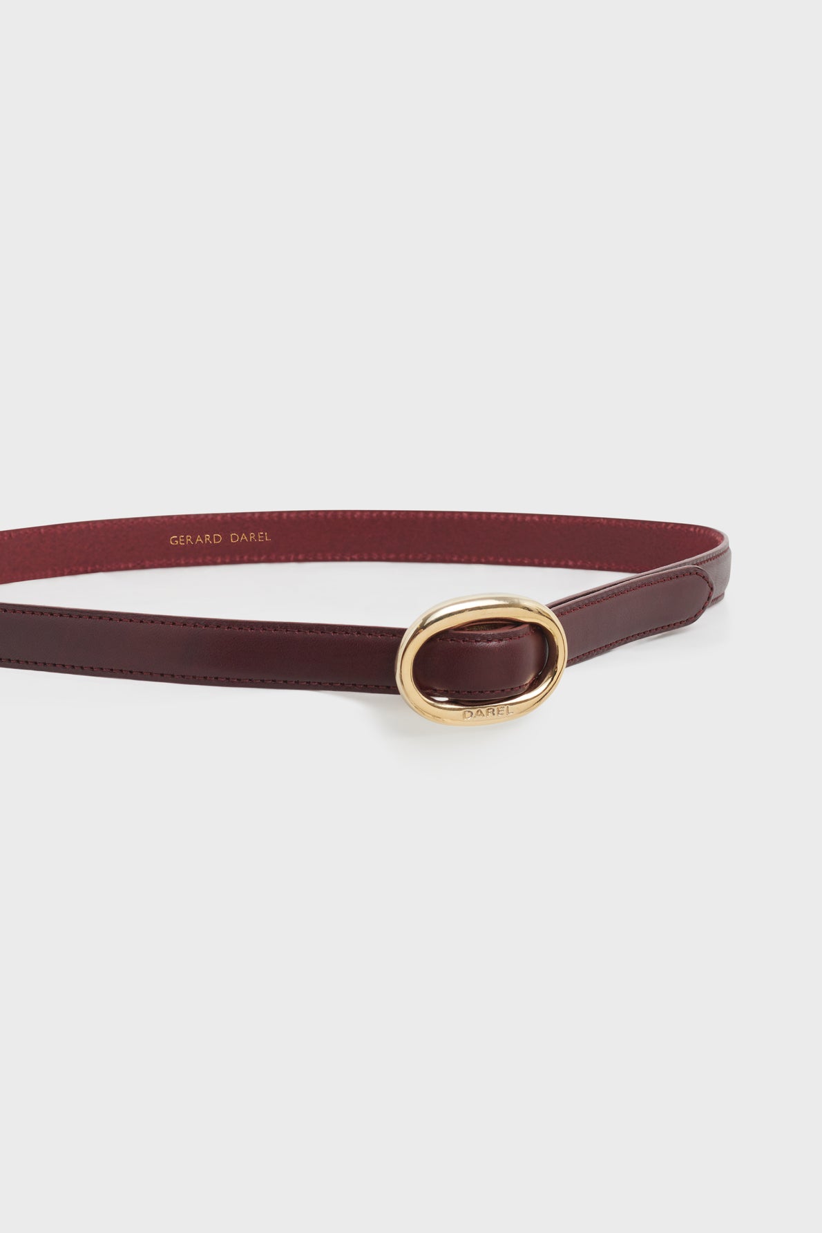 A slim burgundy leather ceinture réglable with a shiny gold oval buckle, displayed on a white background. The inside of this elegant ceinture femme is stamped with the text GERARD DAREL in gold letters.