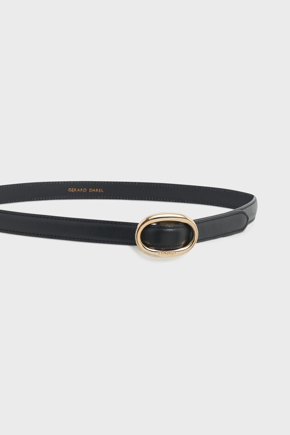 A slim black leather ceinture Sofia with an oval gold buckle, displayed against a plain white background. The brand name Gerard Darel is visible in gold on the inner side of this élégante ceinture femme, making it an essential accessoire de mode for any wardrobe.