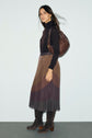 Woman standing against a plain wall, wearing a black turtleneck, a jupe plissée midi with une taille élastiquée, brown suede boots, and carrying a large brown leather shoulder bag.