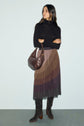 Woman stands against a light background wearing a black long-sleeve top, a jupe plissée ombré brown skirt with une taille élastiquée, dark knee-high boots, and holding a brown shoulder bag.