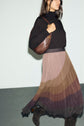 A woman wearing a black turtleneck, a jupe midi plissée with brown gradient tones and a taille élastiquée, dark boots, and carrying a brown shoulder bag stands against a plain light wall.