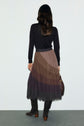 A woman with long brown hair, wearing a black long-sleeve top and a jupe midi à taille élastiquée, stands facing away from the camera against a plain background. Her brown jupe plissée pairs elegantly with knee-high suede boots and a matching brown bag.