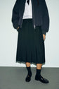 Person wearing a jupe plissée noire mi-longue, white button-up shirt, dark jacket, black socks, and black loafers, standing against a plain background. The look captures an effortless chic à la française. Face is not visible.