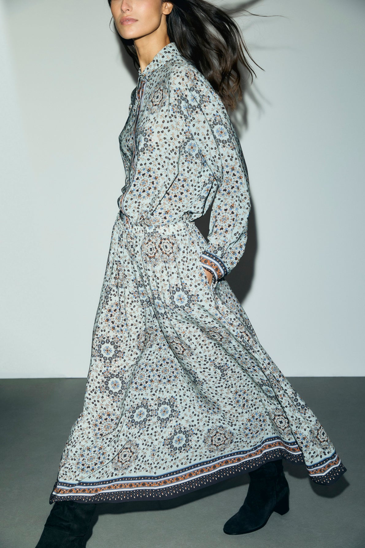 A woman in a patterned long-sleeve maxi dress with a motif floral and black boots walks indoors. Her hair flows behind her, and the dress features a loose fit with geometric patterns, as well as a taille élastiquée for added comfort.