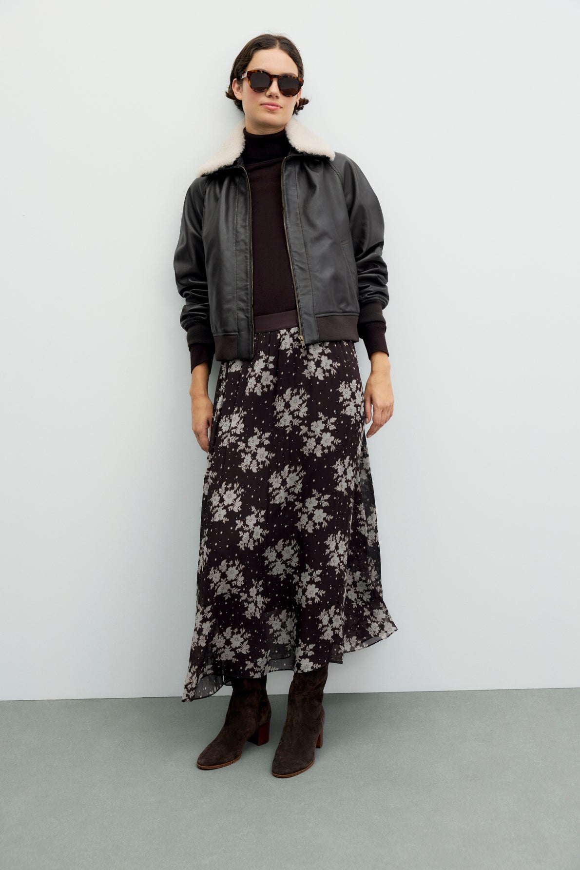 A woman wearing sunglasses, a black leather jacket with a shearling collar, a black turtleneck, and brown boots stands against a plain light background. She completes her look with a jupe en mousseline à imprimé floral de longueur midi.