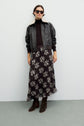 A woman wearing sunglasses, a black leather jacket with a shearling collar, a black turtleneck, and brown boots stands against a plain light background. She completes her look with a jupe en mousseline à imprimé floral de longueur midi.