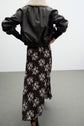 A person stands with hands on hips, wearing a black leather jacket with a white fleece collar over a jupe en mousseline longueur midi à imprimé floral noire and brown boots. The image is taken from behind against a plain background.