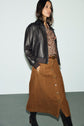 A woman wears a black leather jacket, a paisley blouse, a brown jupe velours trapèze, and black boots, standing against a plain light background.
