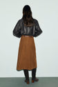 A person with long dark hair stands facing away, wearing a black leather jacket, a brown jupe mi-longue, and brown knee-high boots against a plain light background.