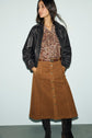 A woman wearing a black leather jacket, paisley print blouse, brown jupe trapèze button-front skirt, and dark knee-high boots stands against a plain light background.