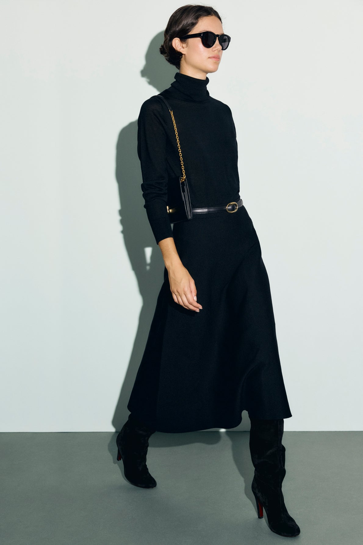 Woman wearing a long-sleeve black maille milano turtleneck dress, black boots, a black belt, black sunglasses, and carrying a small black shoulder bag with a gold chain strap, walking against a light background.