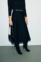 Person wearing a black long-sleeved top, a jupe noire mi-longue in maille milano, black boots, and holding a black handbag with a gold chain strap. The outfit is simple and elegant, complemented by the refined texture of the skirt, and the background is plain.