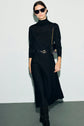 Woman wearing a sleek black turtleneck dress crafted in maille milano, styled with a black belt, black boots, sunglasses, and a black purse with a gold chain strap, standing against a light background.