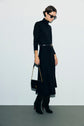 Woman in a black turtleneck dress in maille milano with a belt, knee-high black boots, sunglasses, and a black handbag with a gold chain, standing against a plain light background.