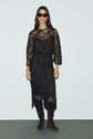 Woman wearing a black dentelle lace dress, dark boots, and black sunglasses stands against a plain light background, facing forward with relaxed posture.