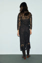 Woman standing with her back to the camera, wearing a black dentelle dress with long sleeves and knee-high dark boots, against a plain light background.