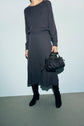 A person wearing a dark maille à côtes sweater and a jupe grise longueur midi, black boots, and holding a black woven handbag stands against a plain light-colored wall. Their face is not visible.
