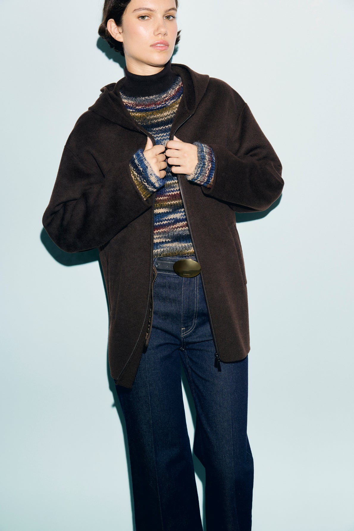A person wearing a dark brown manteau à capuche over a colorful striped sweater and dark blue jeans stands against a light blue background.