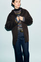 A person wearing a dark brown manteau à capuche over a colorful striped sweater and dark blue jeans stands against a light blue background.