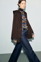 A woman wearing a dark manteau à capuche en laine double face, a multicolored knit sweater, and wide-leg jeans walks against a light gray background.