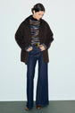 A woman wearing a manteau mi-long dark brown coat in laine double face, a striped turtleneck sweater, wide-leg blue jeans, and brown shoes stands against a plain light background, looking down.