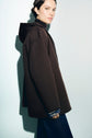 Woman wearing a dark brown manteau à capuche with blue and gray sweater cuffs, standing sideways against a pale background and looking toward the camera.