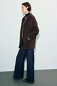 Person stands in profile wearing a dark brown manteau mi-long, striped sweater, wide-leg dark blue jeans, and brown shoes against a light background. Hands are in jacket pockets.