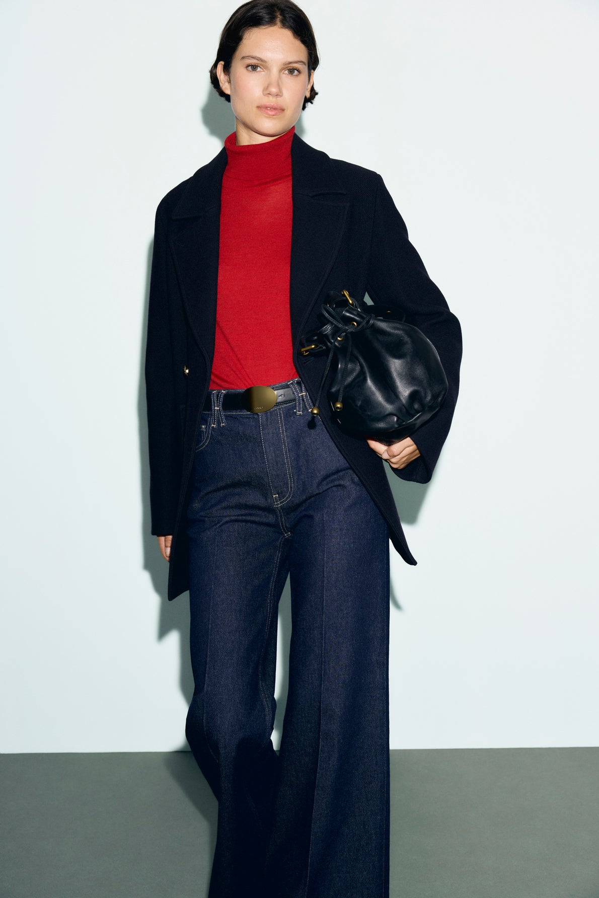 A person wearing a red turtleneck, a caban bleu marine over a dark blazer, wide-leg jeans, and holding a black bag stands against a light background.