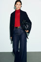A person wearing a red turtleneck, a caban bleu marine over a dark blazer, wide-leg jeans, and holding a black bag stands against a light background.