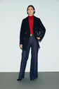 A person wearing a black manteau mi-long, red turtleneck, blue flared jeans, and black boots holds a small black bag, standing against a plain light background.