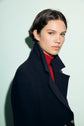 A person with short dark hair wears a caban bleu marine over a red turtleneck, looking at the camera with a neutral expression against a pale background.