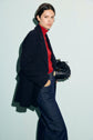 A person in a black blazer, red turtleneck, and dark jeans stands against a light background, holding a black handbag crafted from laine vierge and looking slightly past the camera with a neutral expression.