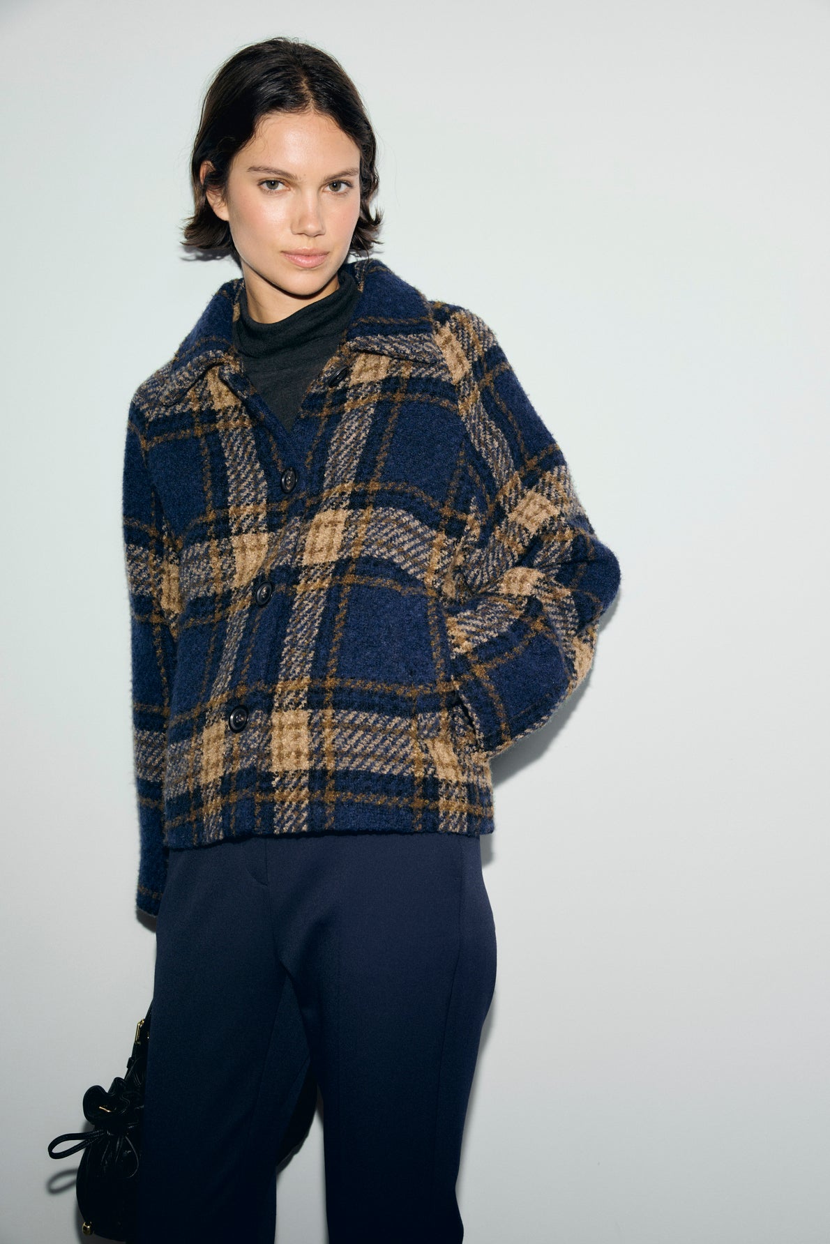 Woman standing against a plain wall, wearing a blue and tan manteau à carreaux over a dark turtleneck and navy pants, with one hand in her pocket and a neutral expression.
