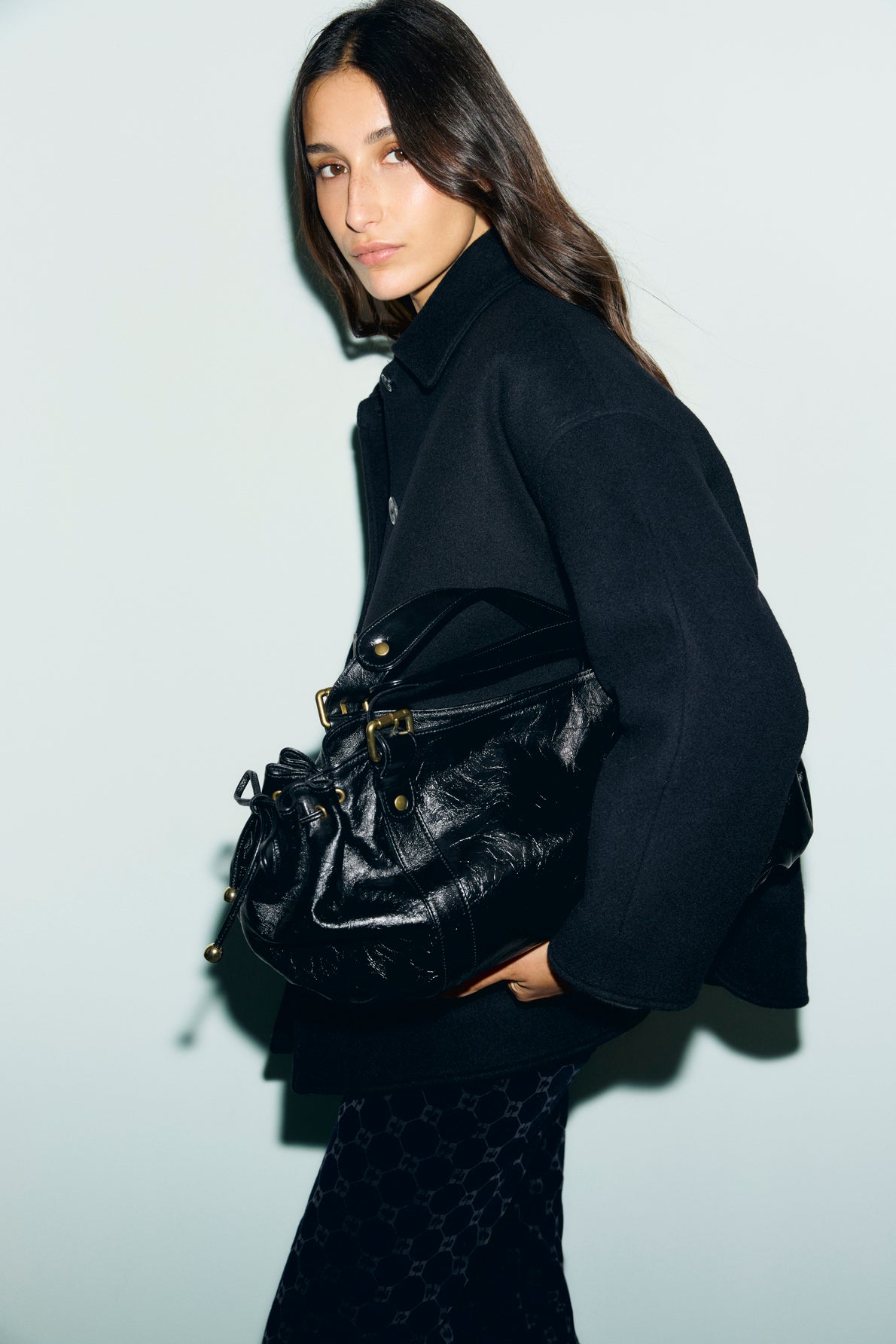 A woman with long brown hair, wearing a manteau court in black paired with patterned black pants, stands holding a large black leather handbag with gold accents against a plain light background.