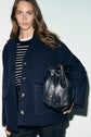 Woman with long brown hair wears a manteau chic femme bleu marine over a black and white striped shirt, holding a black leather drawstring bag. She stands against a pale background and looks into the camera.