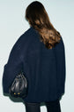 A person with long brown hair, seen from behind, wears a manteau bleu marine oversized coat and dark pants, holding a small black drawstring bag with gold accents.