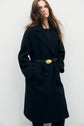 A woman with long brown hair wears a chic à la française black belted manteau long in drap de laine with large lapels, standing against a plain white background with her hands in the coat pockets.