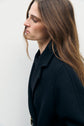 A person with long brown hair, wearing a manteau long in drap de laine, looks downward and to the side against a plain light background, embodying un chic à la française.