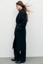 Woman in a manteau long made of drap de laine with a belt, wide-leg pants, and black shoes stands sideways against a plain white wall; her long hair flows back as if blown by wind, embodying un chic à la française.